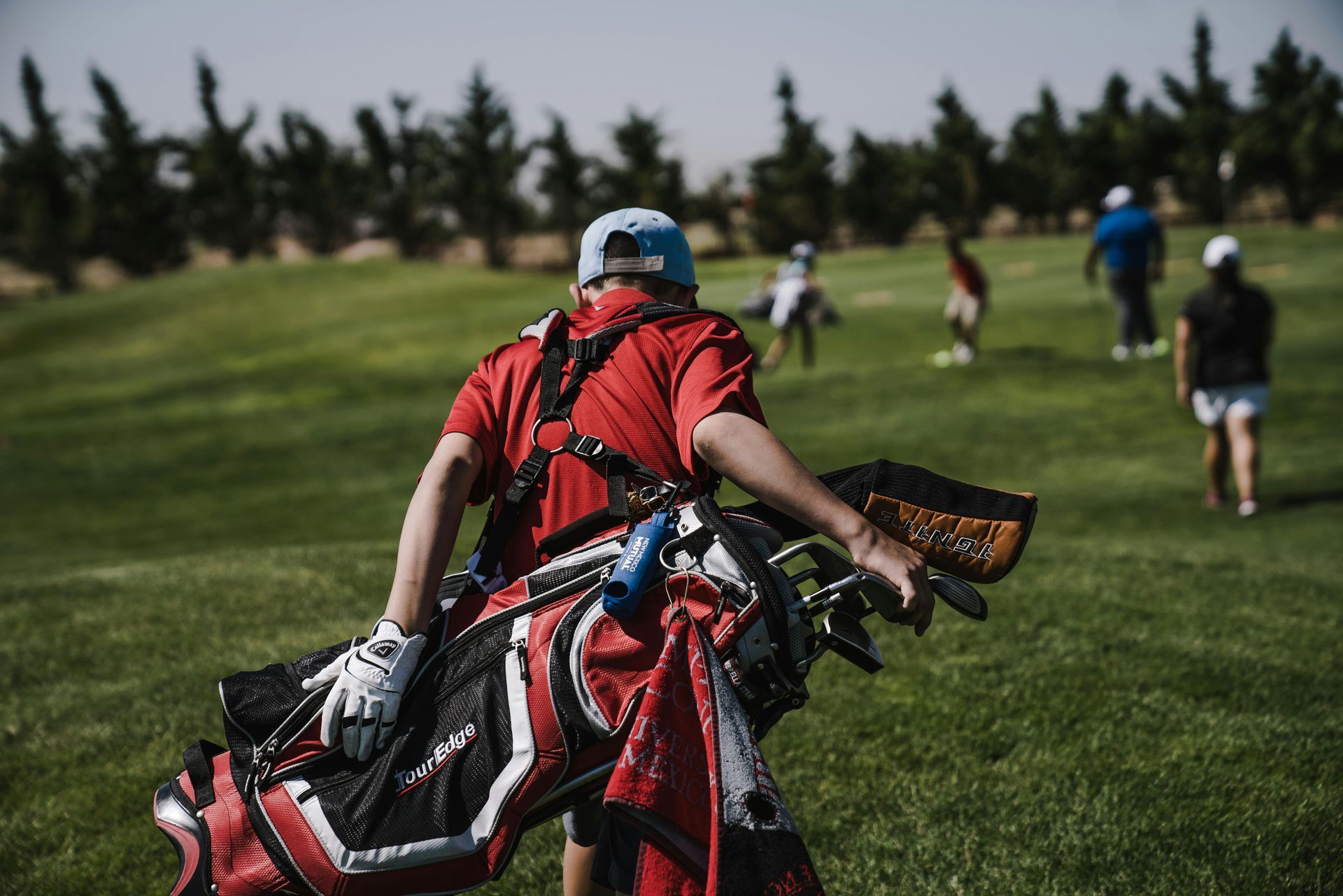 Person carrying a golf bag on a green course, with other golfers visible in the background. The person wears a red shirt and a blue cap.
