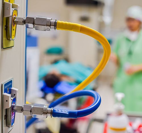 Oxygen and air hoses connected to wall outlets in a hospital room. Patient in background.