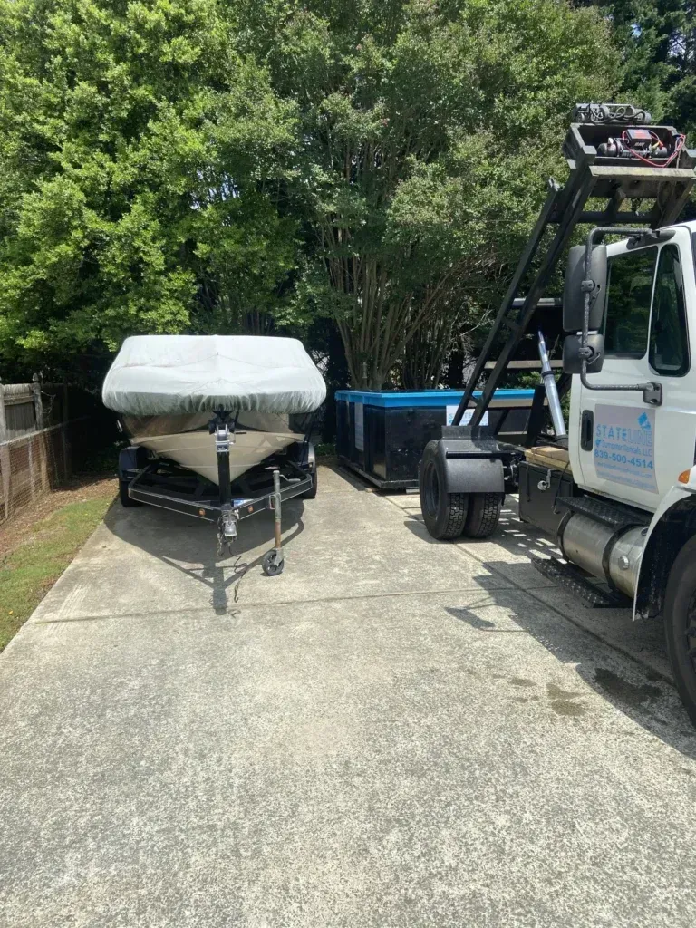 Boat on a trailer next to a hot tub, parked on a concrete driveway beside a large truck and green trees.