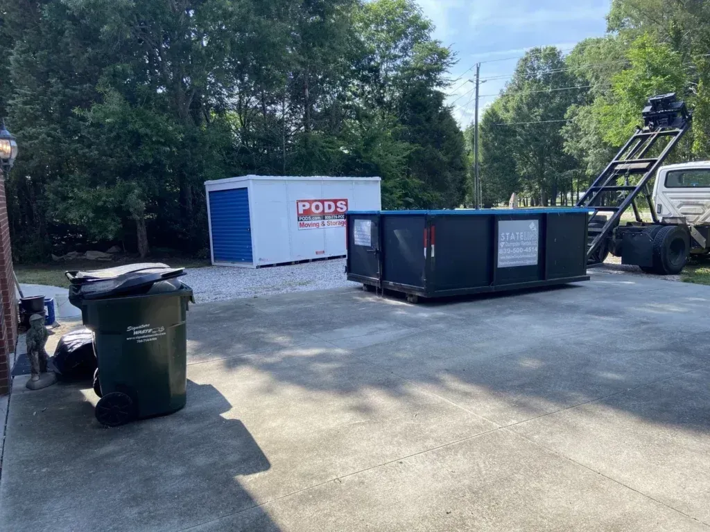 A dumpster, storage unit, and trash cans on a paved lot. Trees and a truck are in the background.