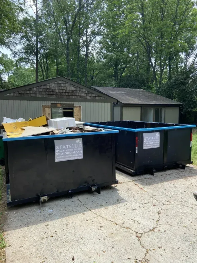 Two black dumpsters sit in front of a house undergoing renovation, with a debris pile visible.