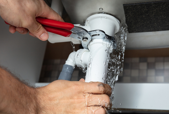 Hands using pliers to tighten a leaking white pipe under a sink, with water spraying out.