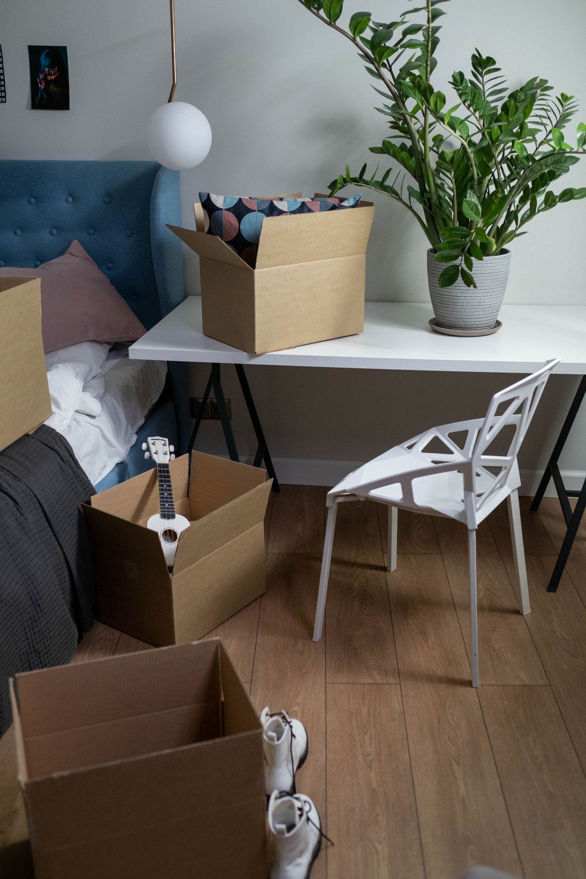 Moving boxes in a bedroom with a white desk, chair, bed, and a potted plant