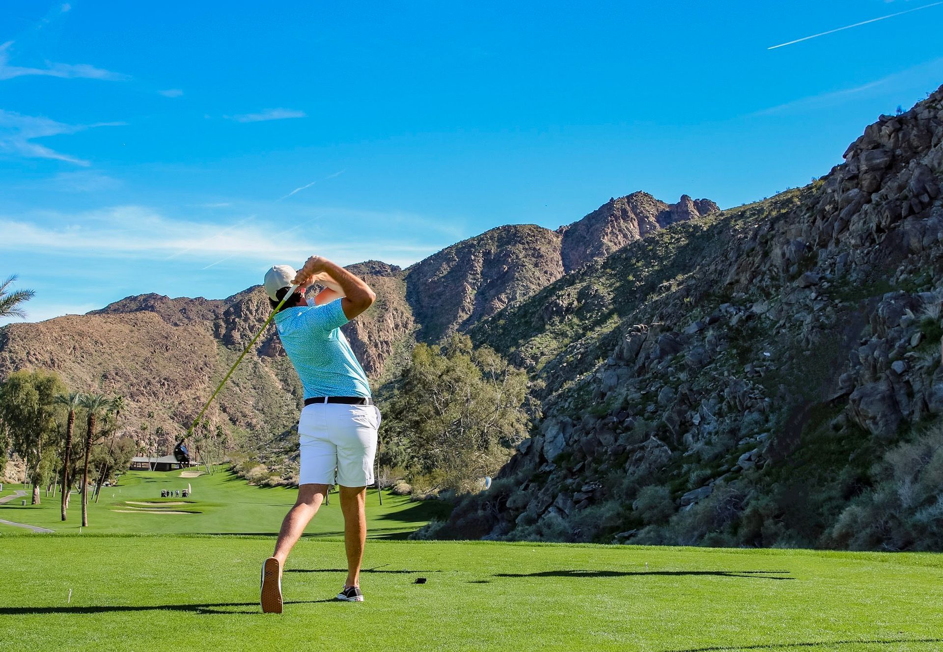 Golfer swinging on green, mountain backdrop, sunny sky.