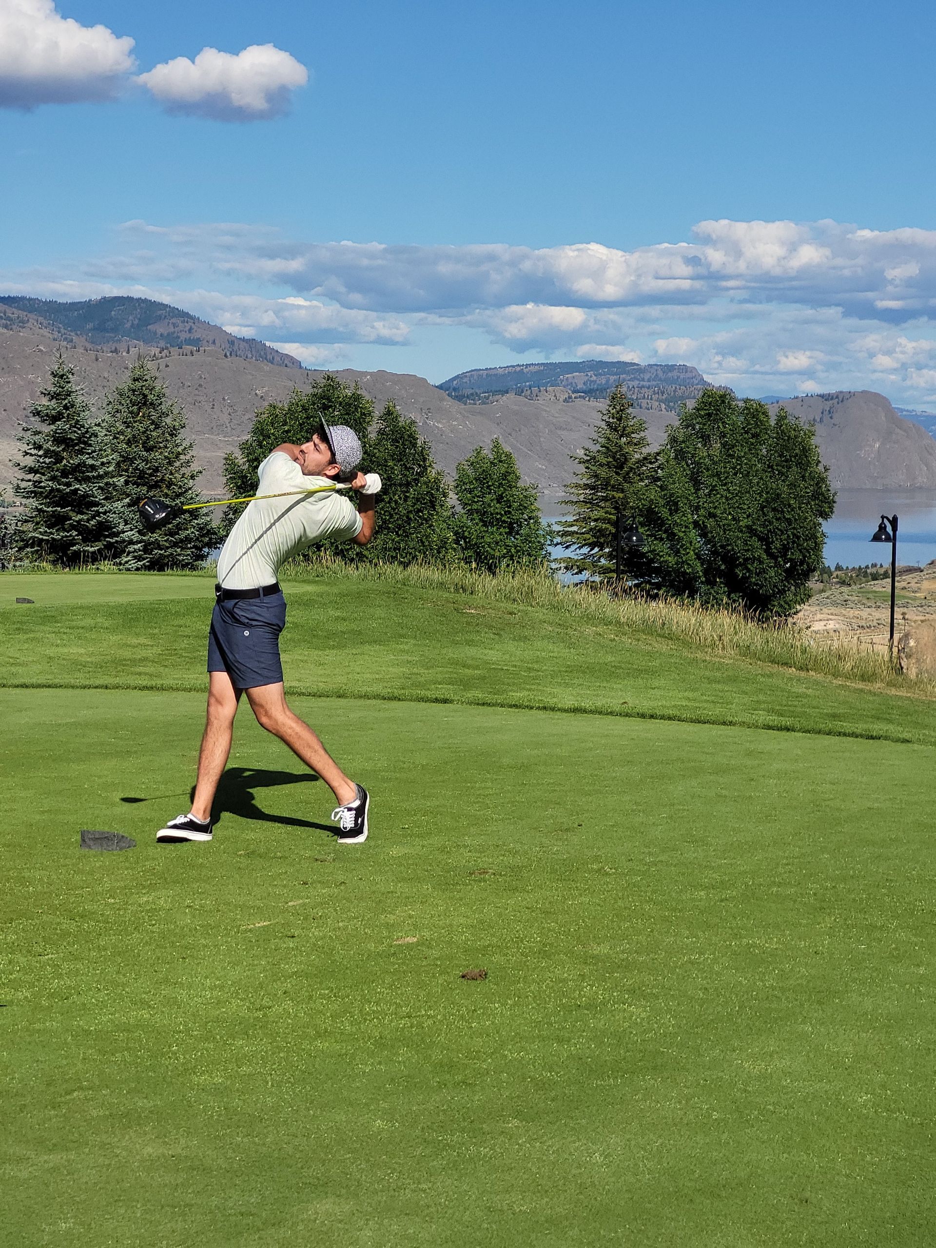 Golfer mid-swing on a green golf course, mountains and lake in background. Sunny, blue sky.
