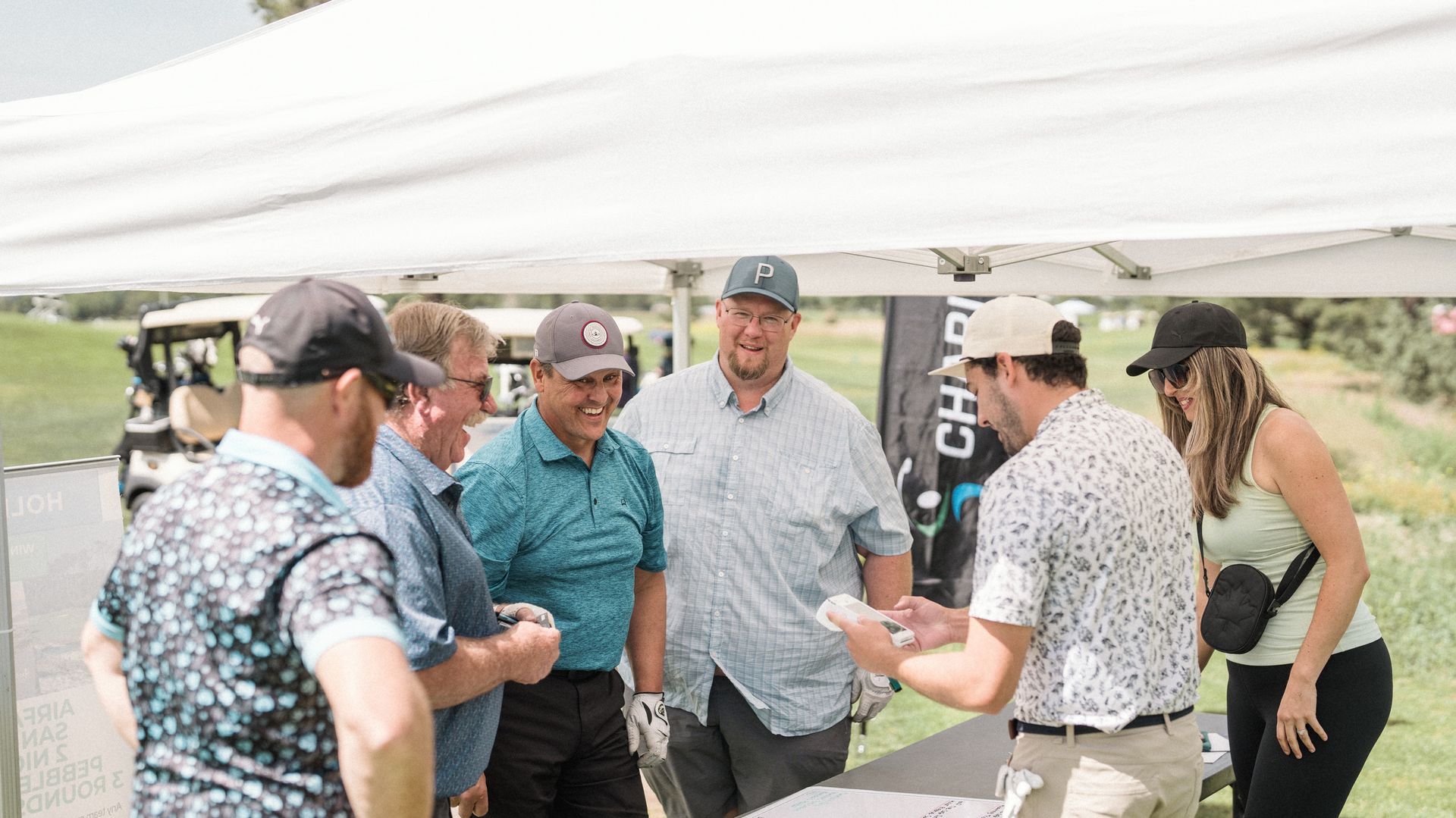 People gathered under a white tent at a golf event, looking at materials held by a person.