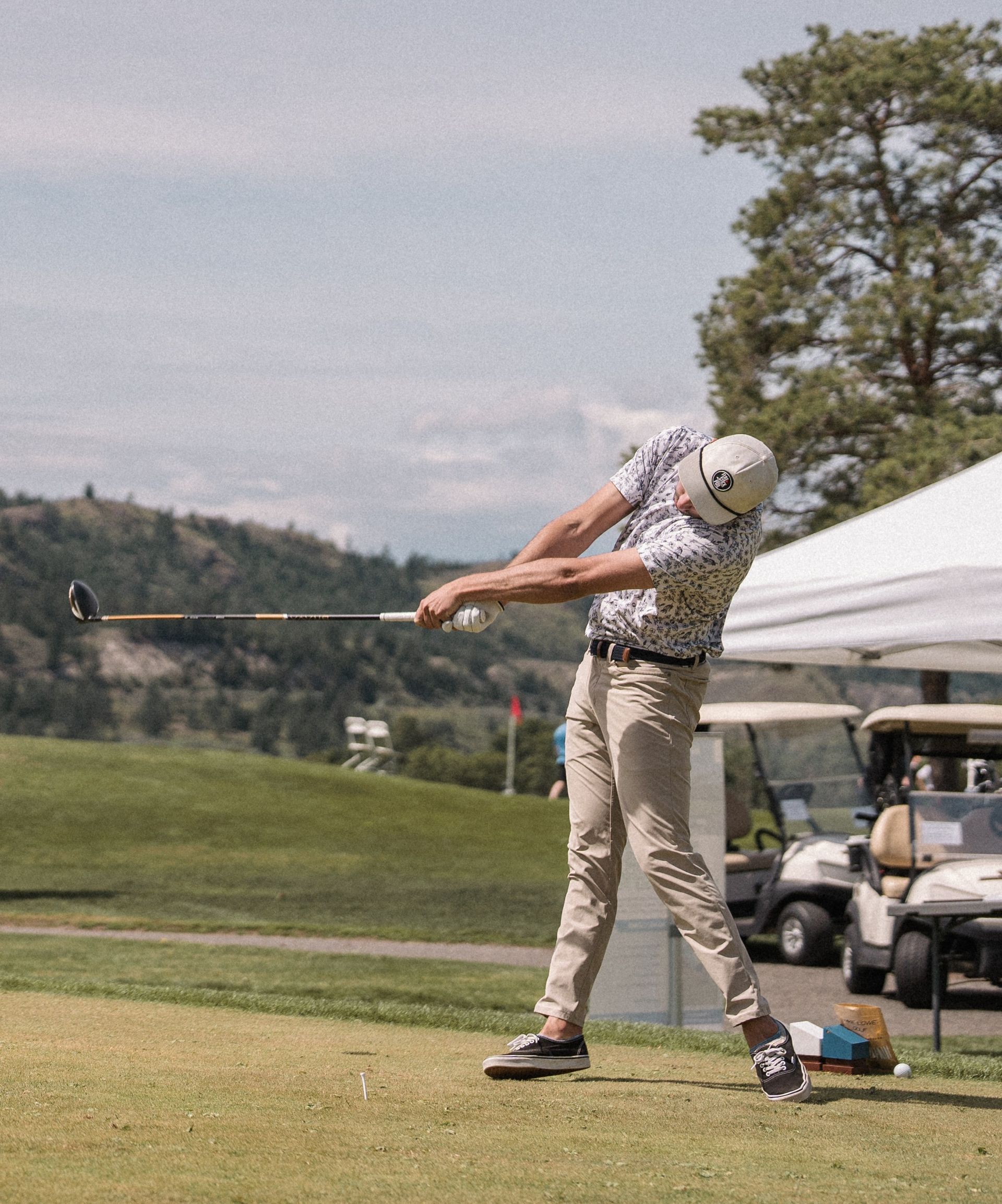 Golfer swings a club on a green course. A tent and golf carts are in the background, with hills and blue sky.