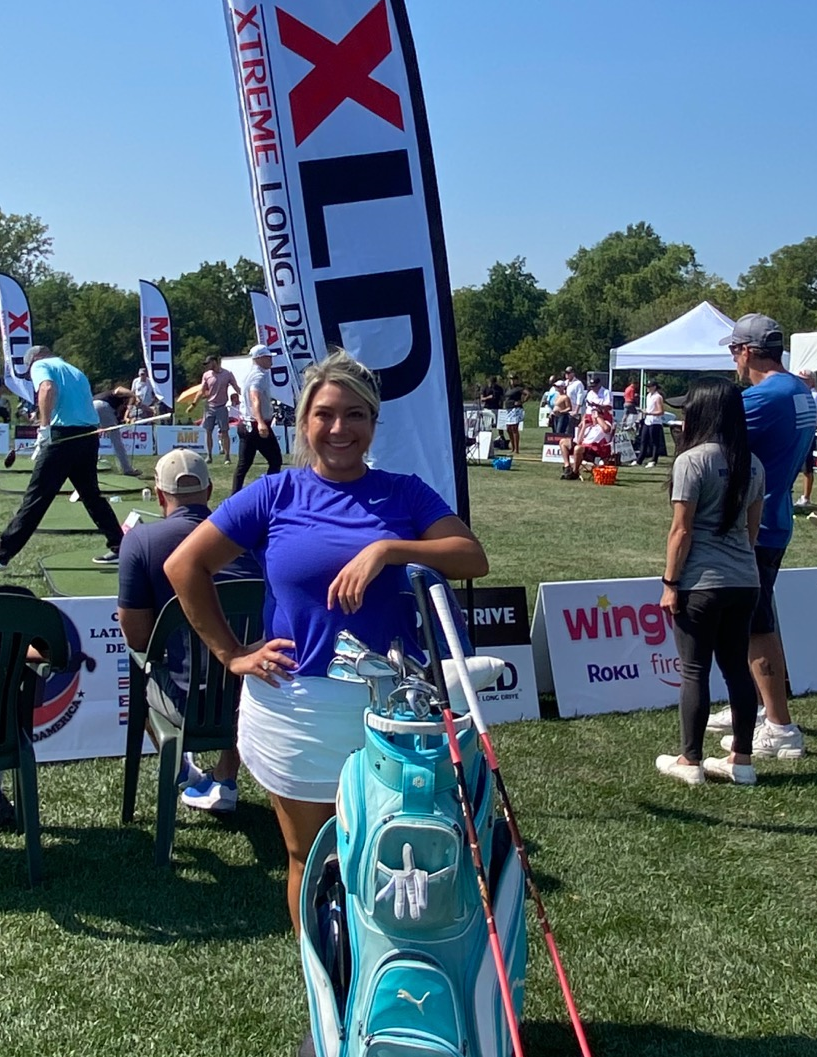 A woman is standing in front of a xld banner holding a golf bag.
