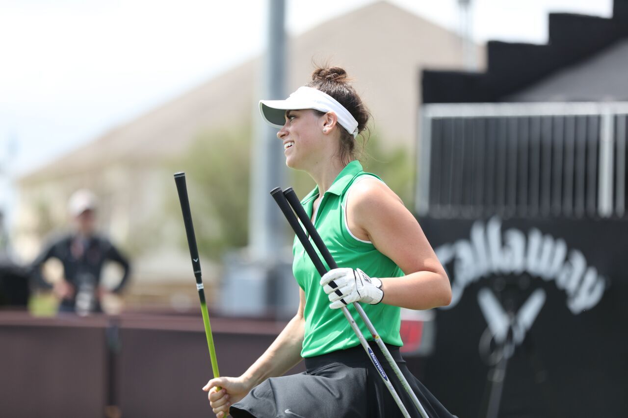 A woman in a green shirt and white visor is holding two golf clubs.
