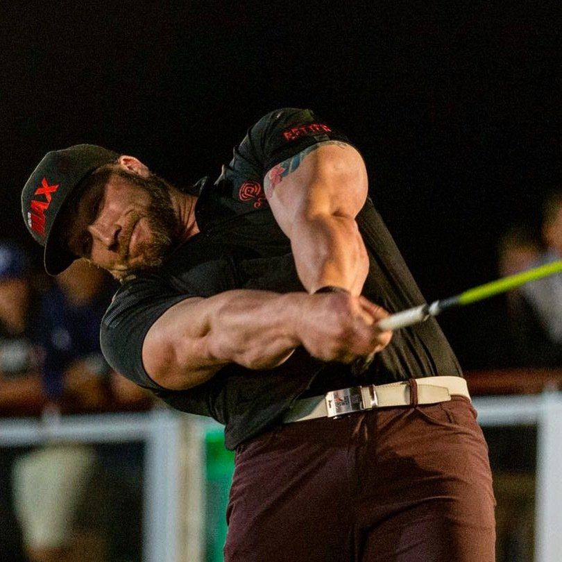 Man in black shirt and cap swings golf club, large biceps flexed, brown pants, outdoor setting.