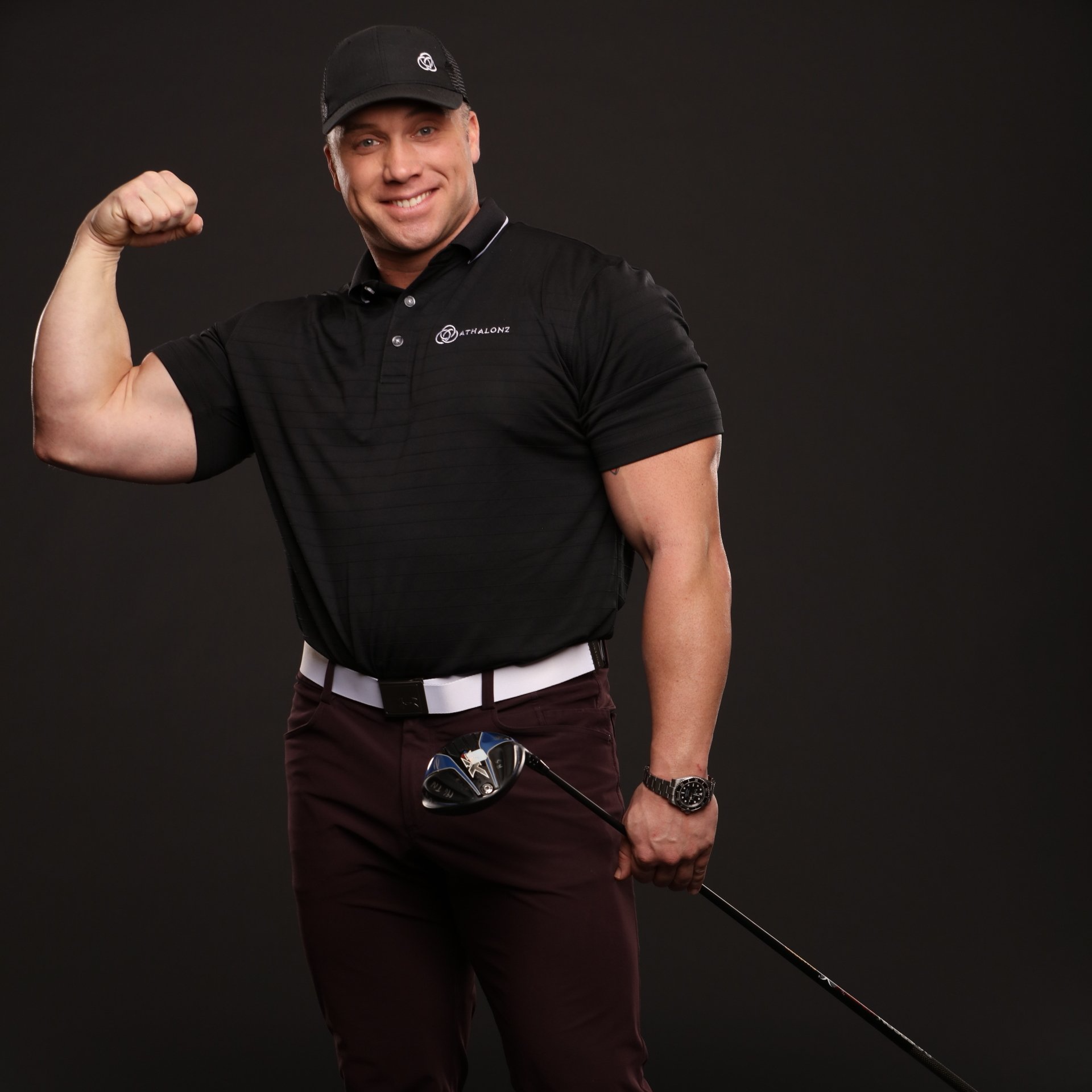Man flexing bicep, holding golf club. He's wearing a black polo shirt, dark pants, and a baseball cap. Dark background.