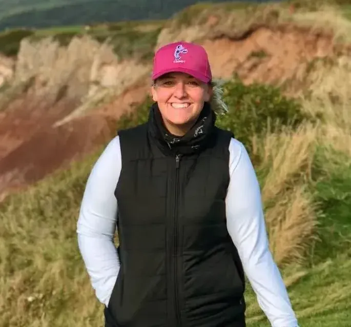 Woman wearing a pink cap and black vest smiles outdoors near a coastal bluff.