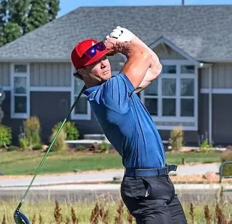 Golfer in red cap swings club in front of a house, blue shirt, black pants.