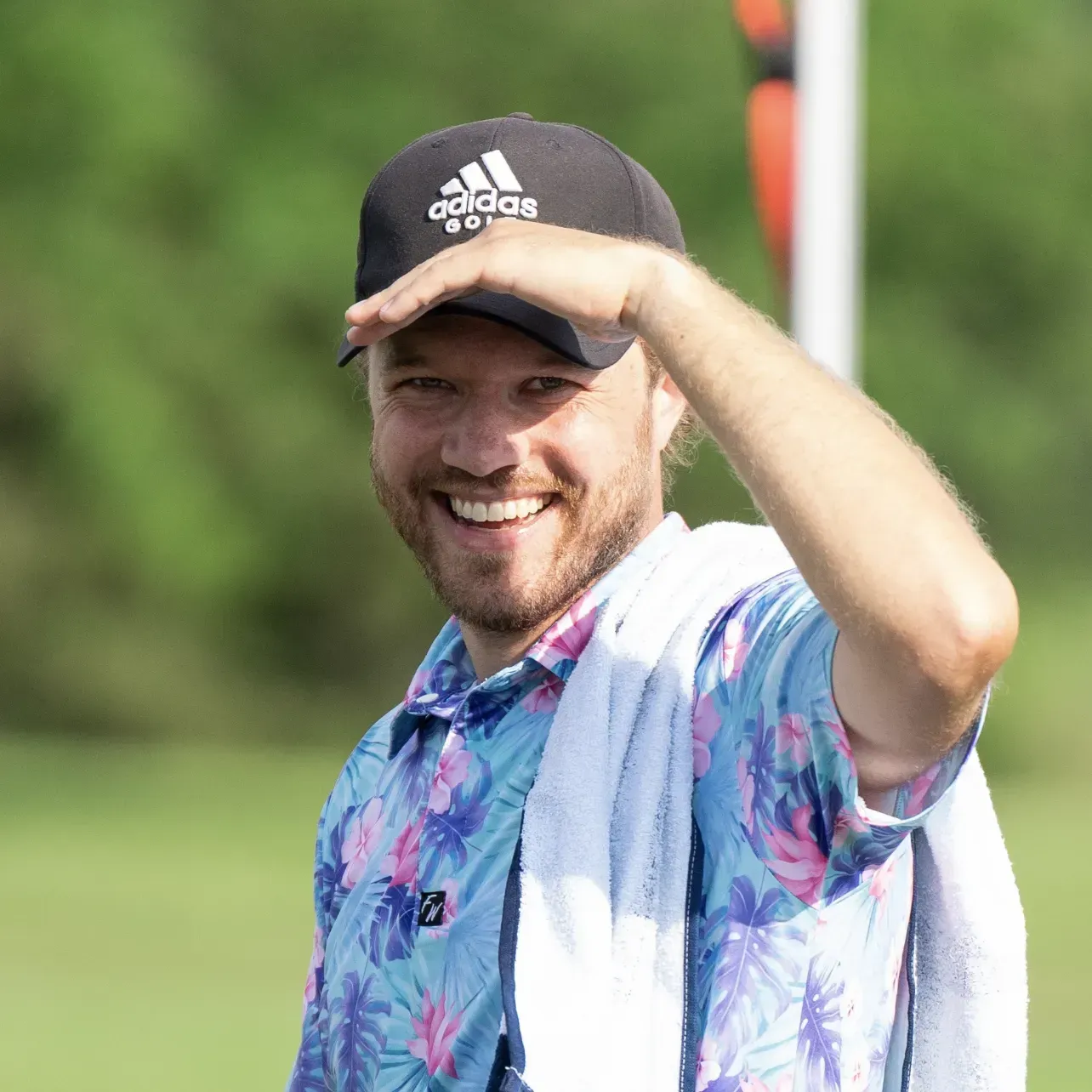 Man in floral shirt and Adidas hat shielding eyes, smiling on a golf course.