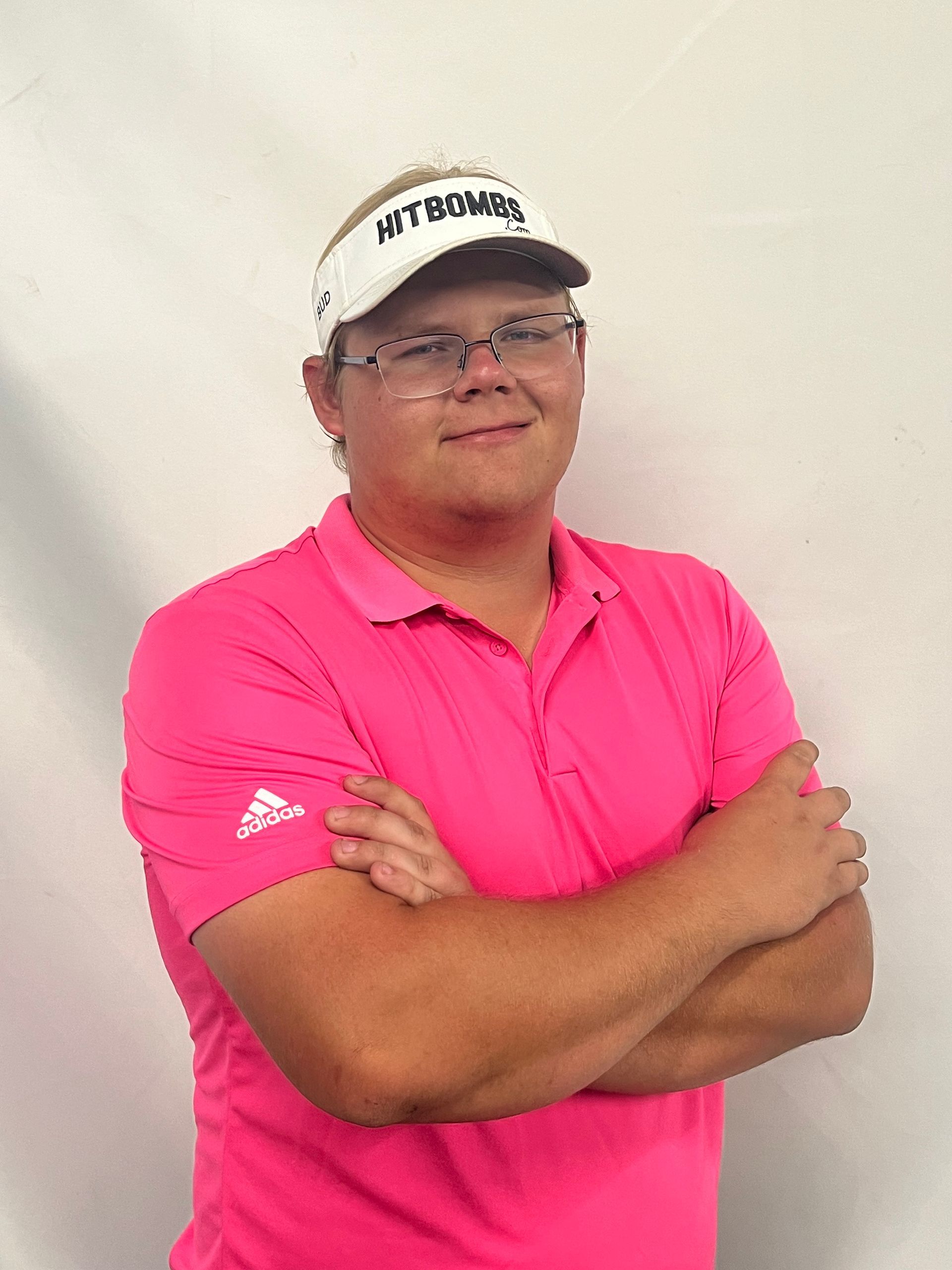 A man wearing a pink shirt and a white hat is standing with his arms crossed.