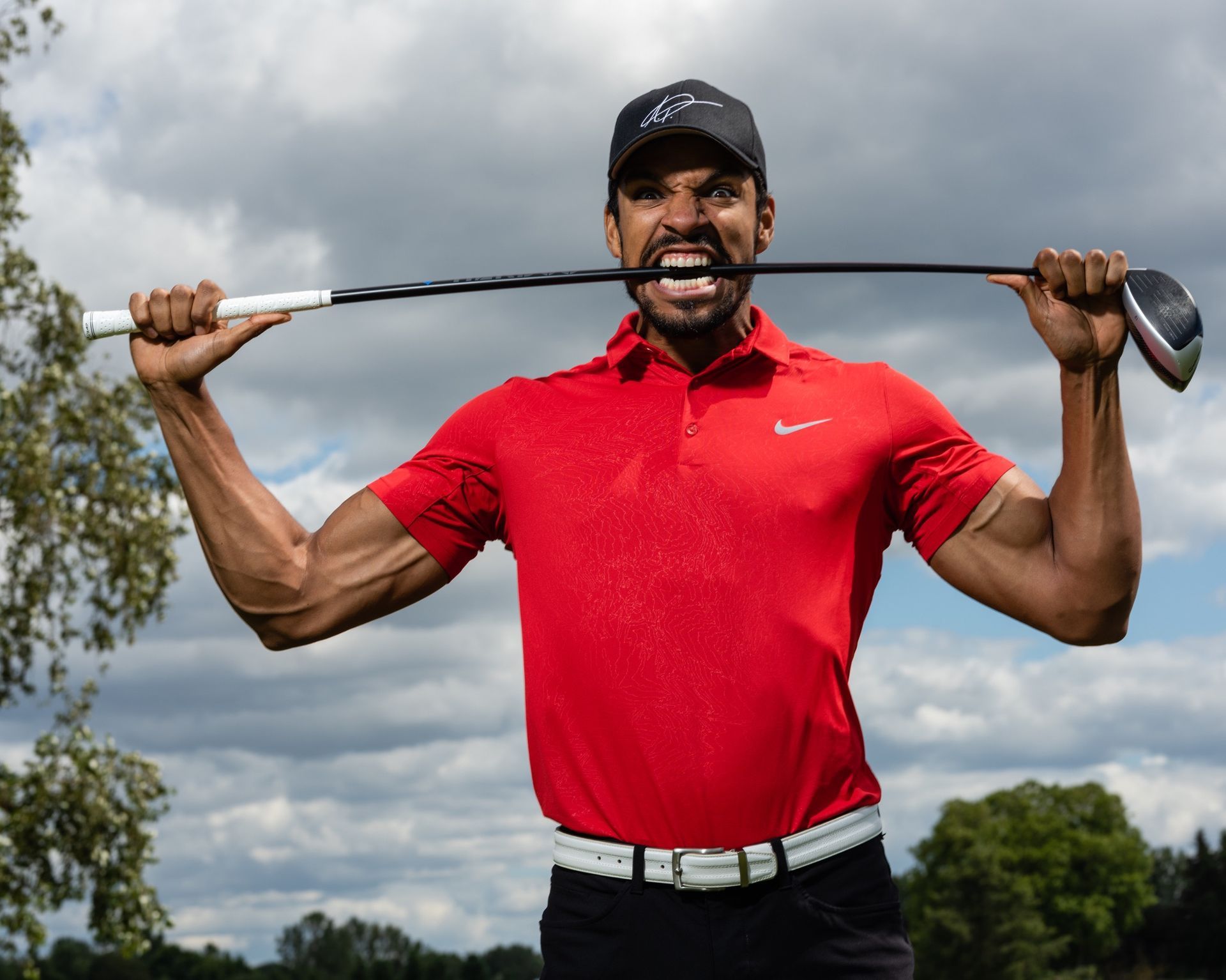 Man holding a golf club in his mouth, wearing a red shirt, white belt, and black hat; outdoors on a cloudy day.