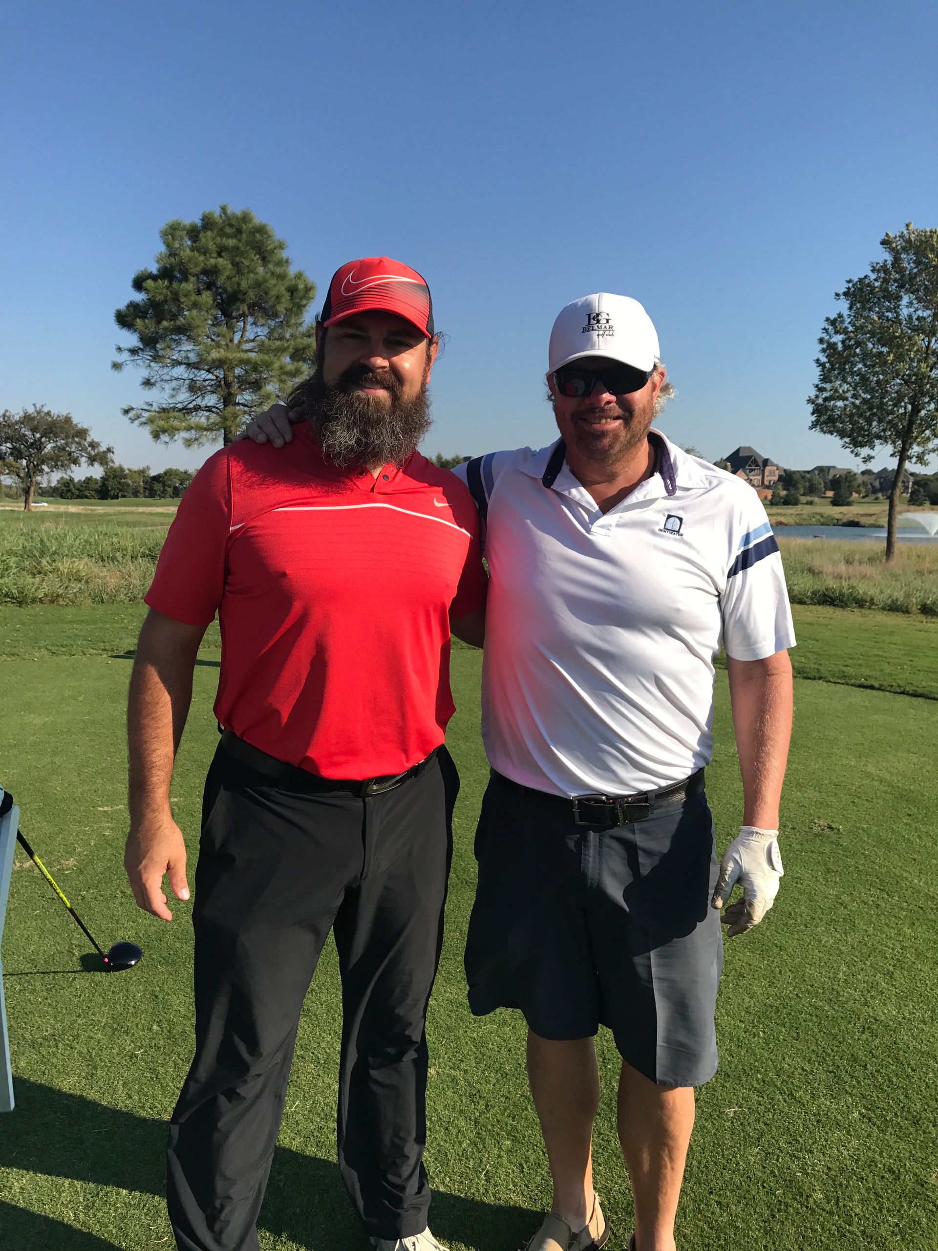 Two men on a golf course, one in red shirt, beard, other in white shirt, arm around each other.