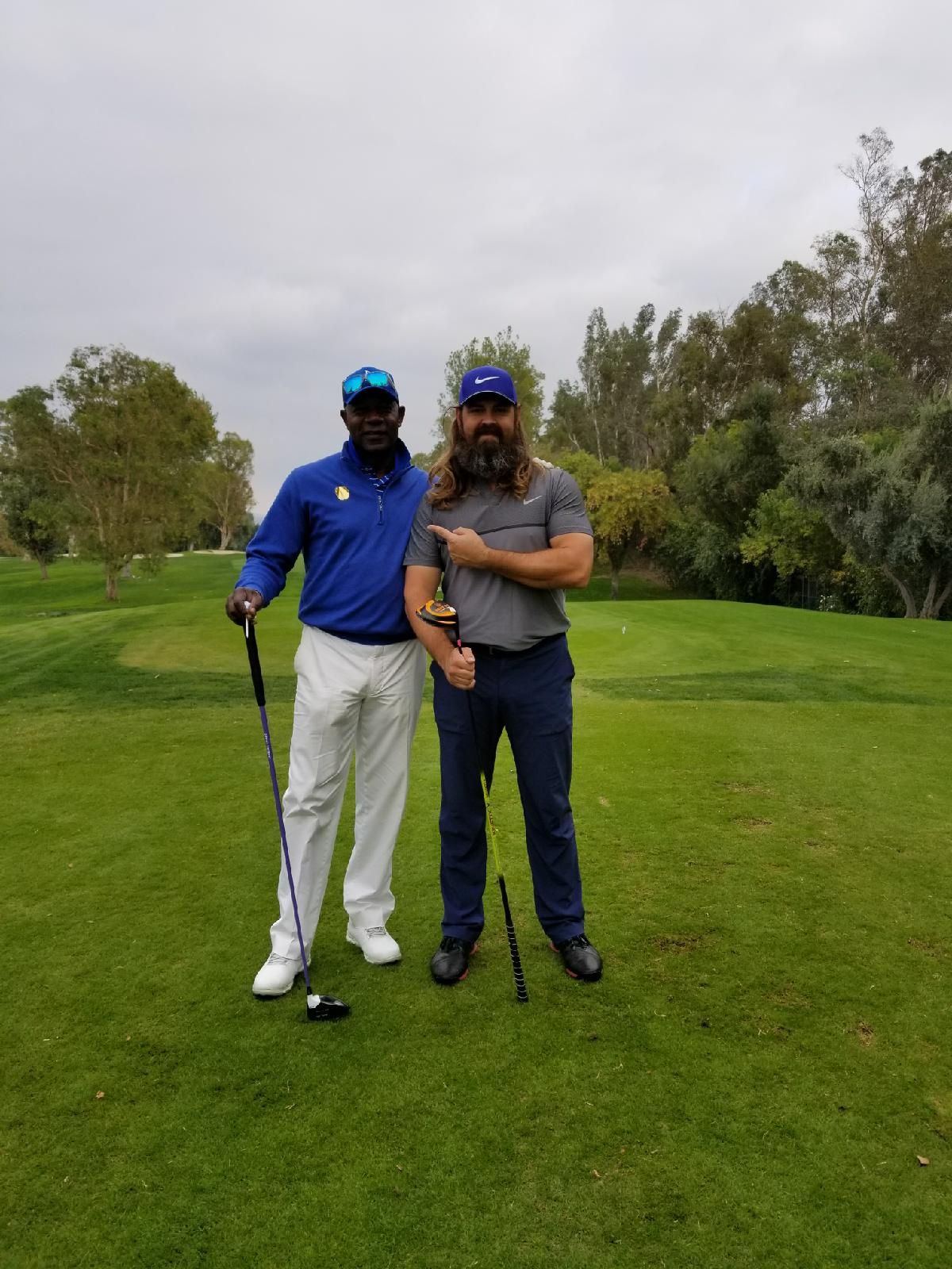 Two men on a golf course, one points to his golf club. Overcast day.