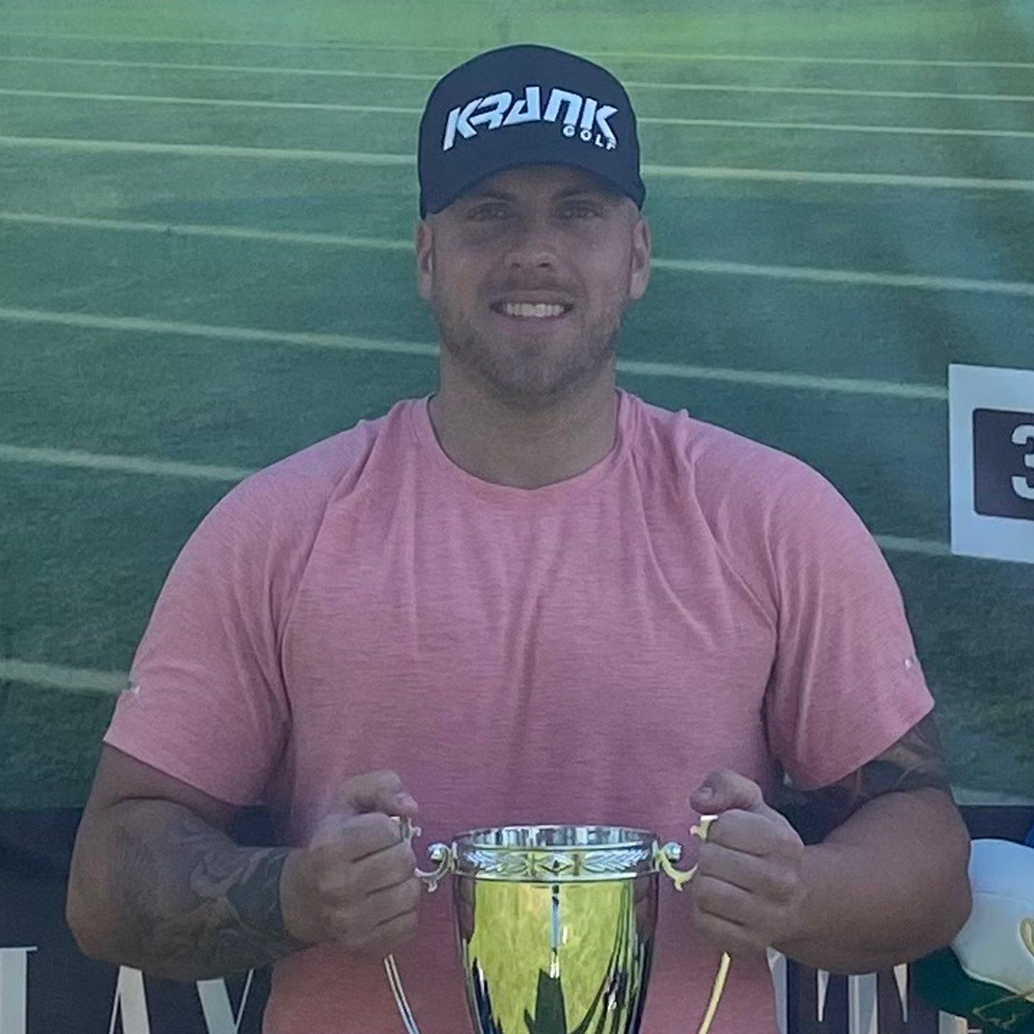 A man wearing a pink shirt and a black hat is holding a trophy.