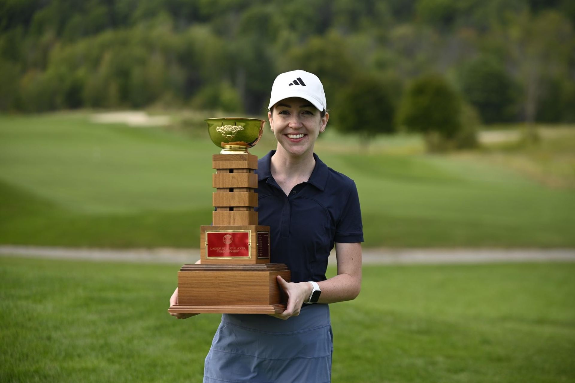 Woman in golf attire holding trophy on a golf course. She is smiling.