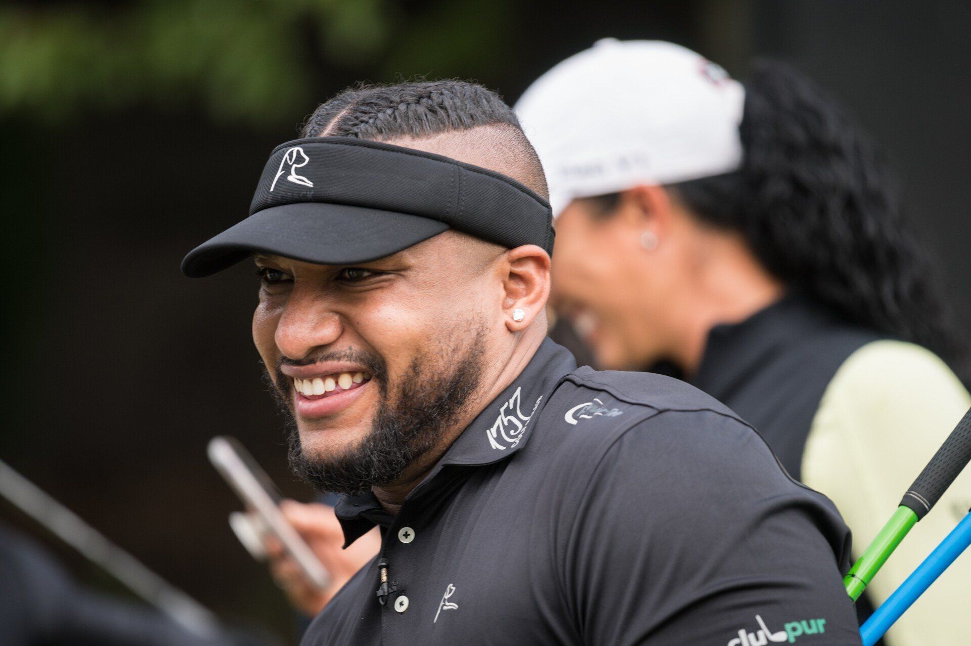 Man smiling, wearing a black visor and shirt with logo, holding golf club, outdoors.