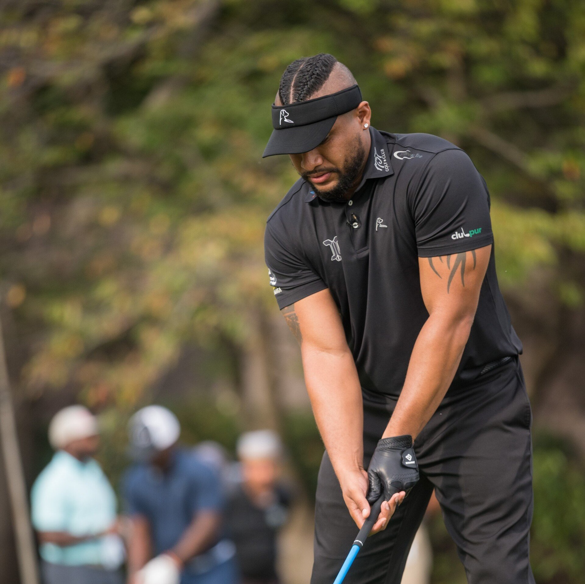 Man in black golf attire, visor, preparing to swing club on a course.
