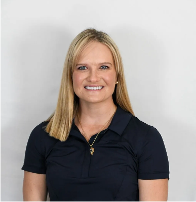 Woman with blonde hair wearing a dark blue polo shirt smiles in front of a white background.