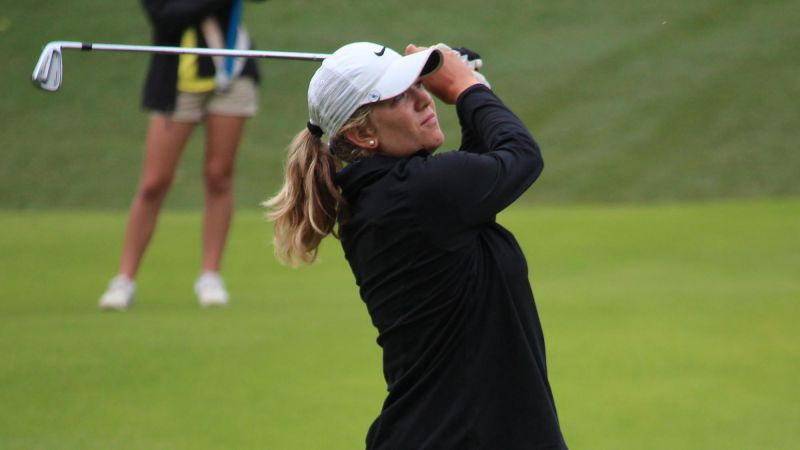 Golfer in black jacket swings club on green course, wearing a white hat.