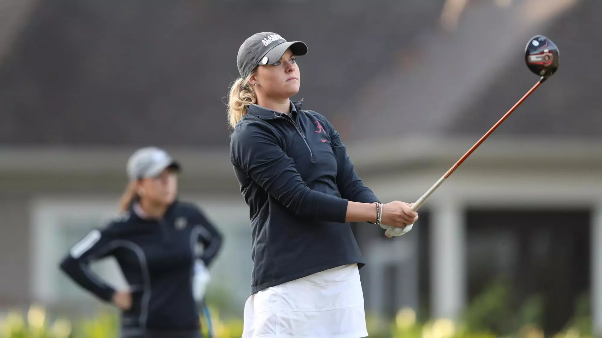 Woman golfer swinging club on golf course; another woman watches in background.