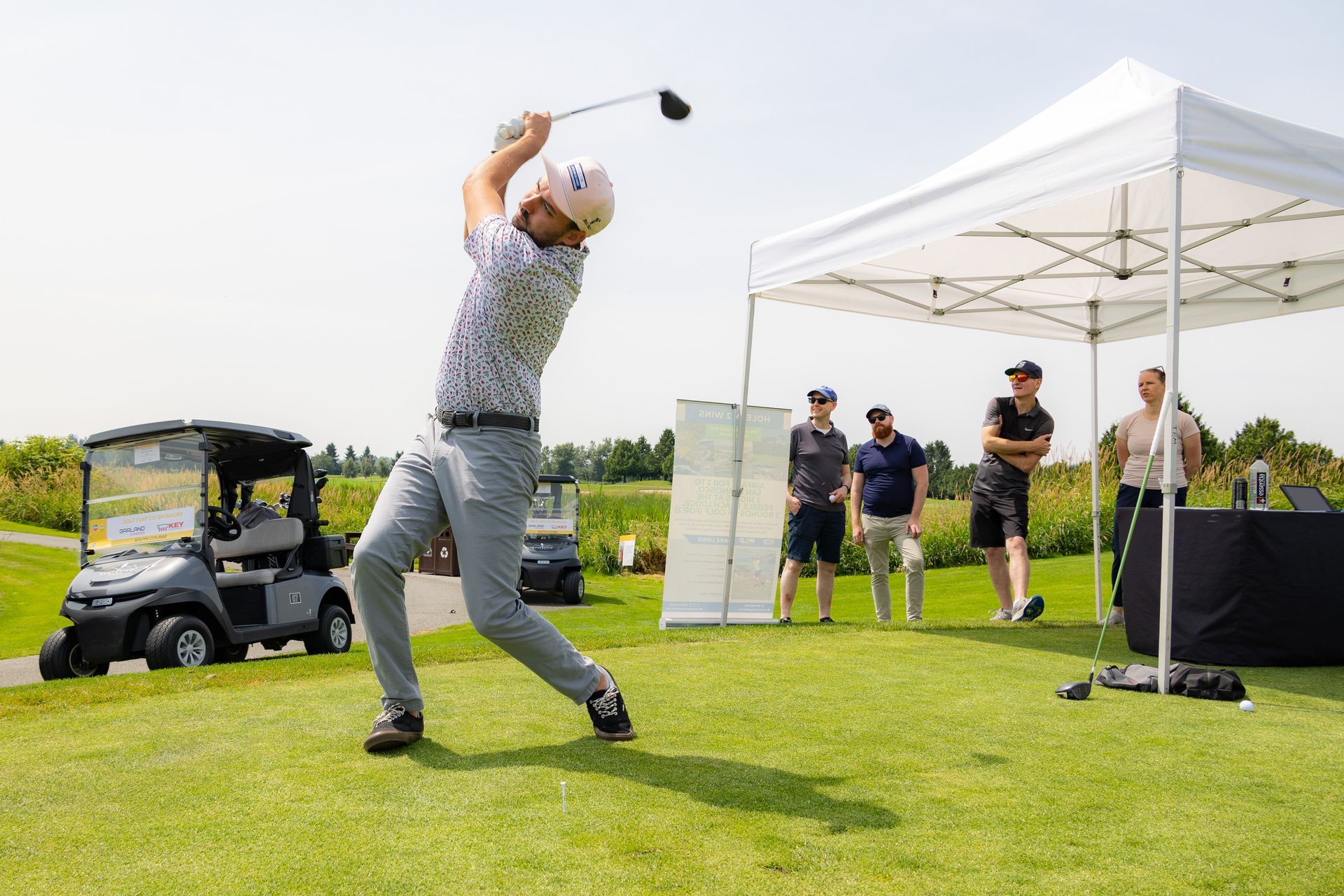 Golfer swings club on green; spectators watch under a white tent; golf cart in background.