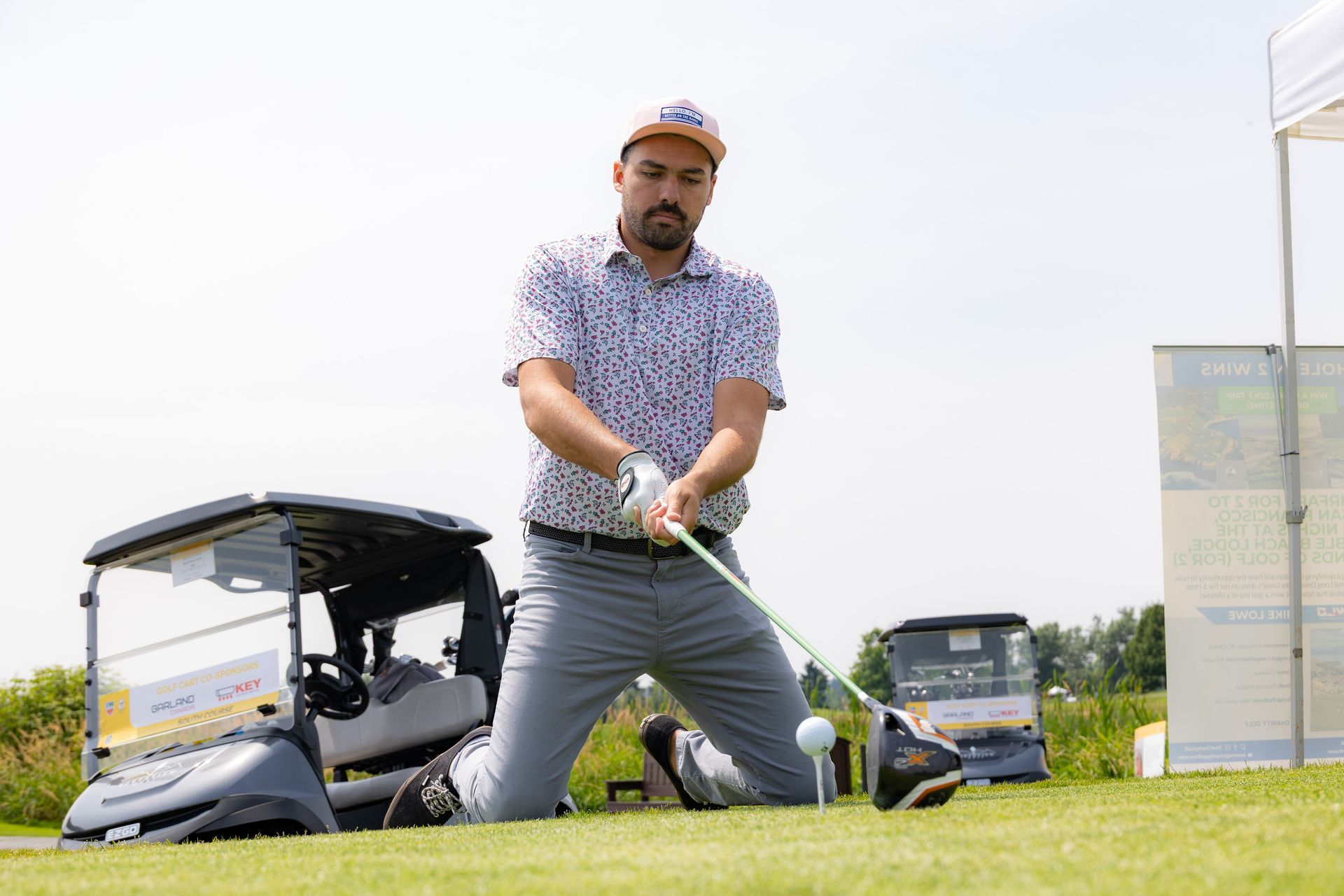 Man kneeling on golf course, about to swing club toward a golf ball; golf cart in background.