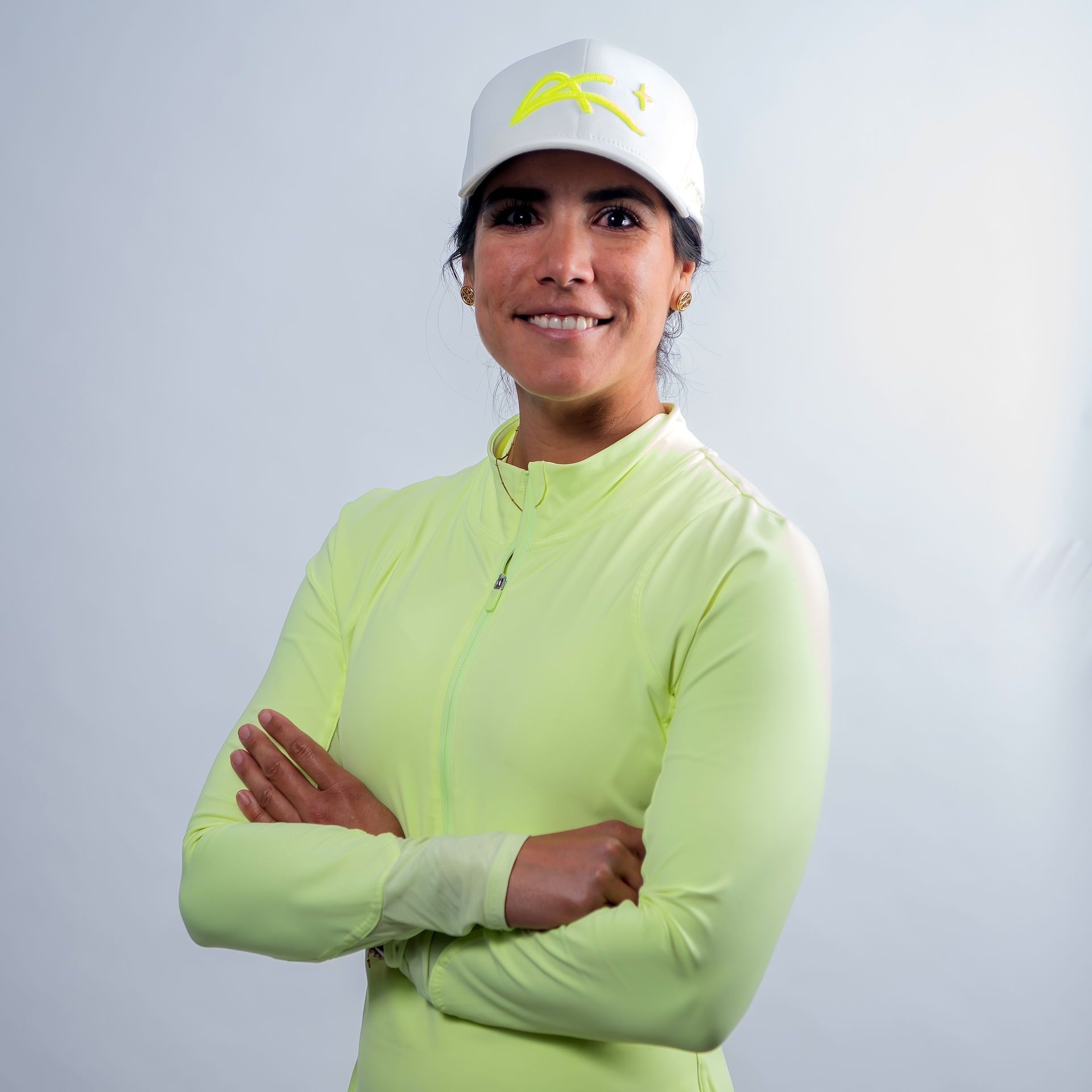 A woman wearing a yellow shirt and a white hat is standing with her arms crossed.