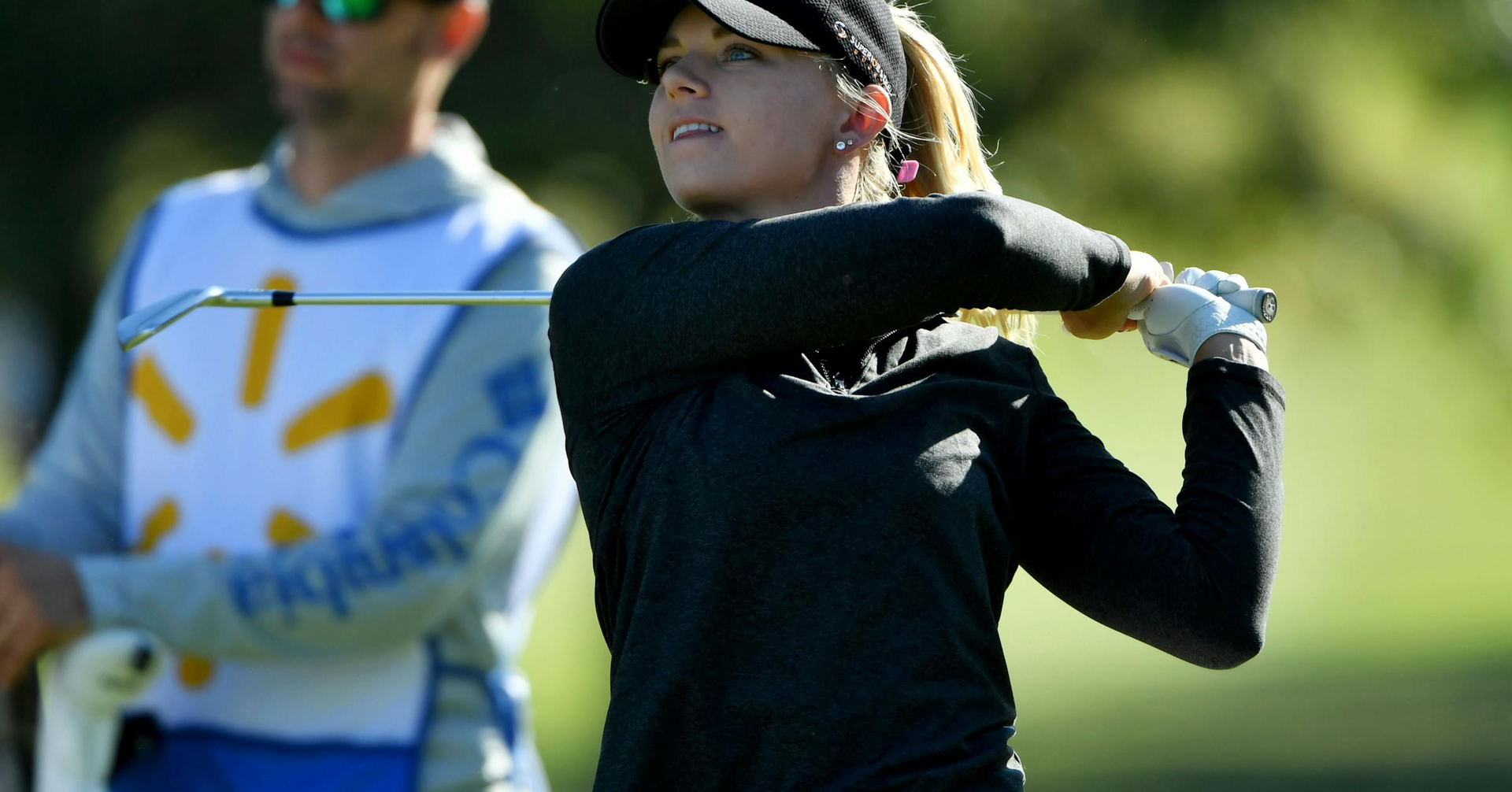 Woman in black swinging a golf club, golf course in background, caddy nearby.