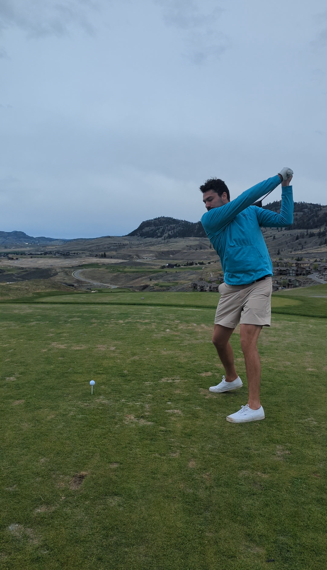 Person in a blue jacket swinging a golf club on a green course with a cloudy sky and mountains in the background.