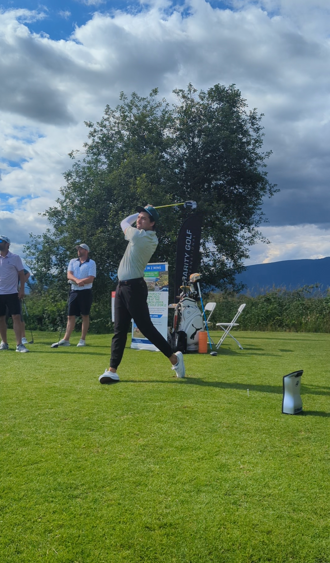 Golfer swinging club on a green course, blue sky with clouds, trees and spectators in background.