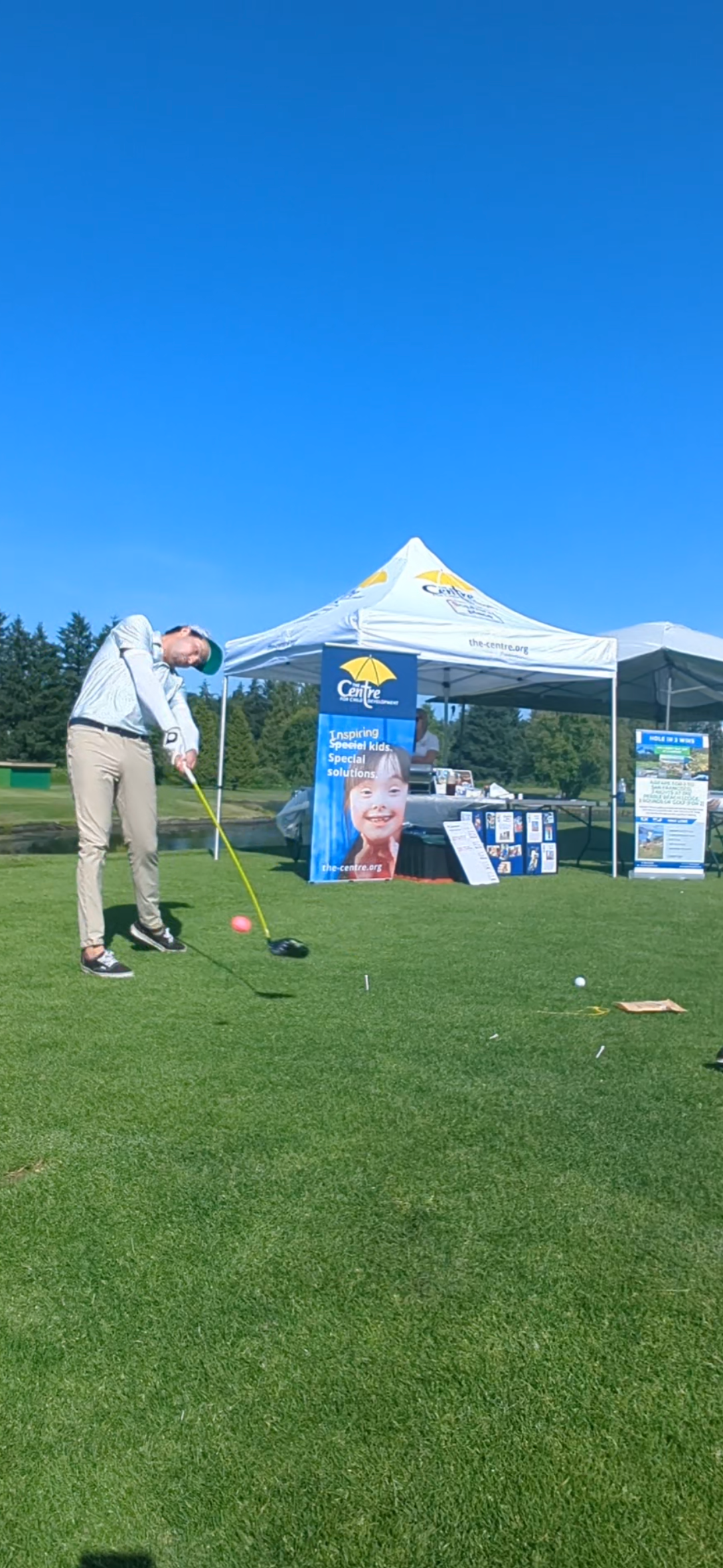 Person swings golf club on a green field under a clear blue sky, tent in the background.