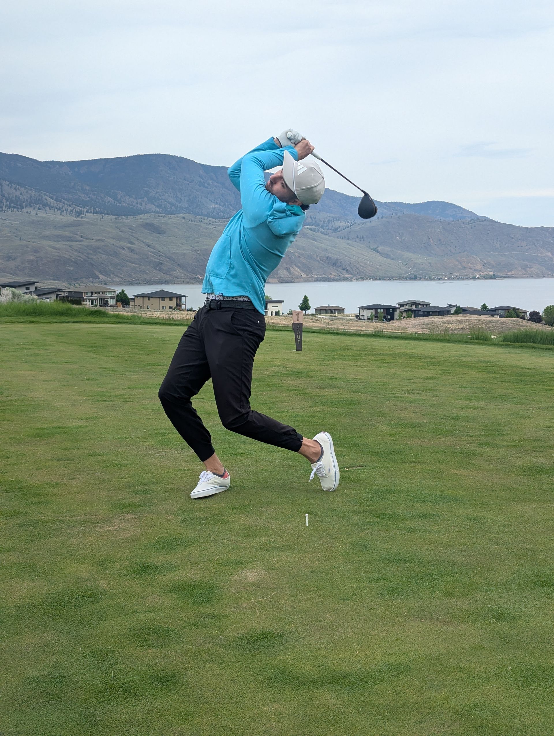 Golfer in a blue shirt and black pants mid-swing on a golf course. Mountains and water in the background.