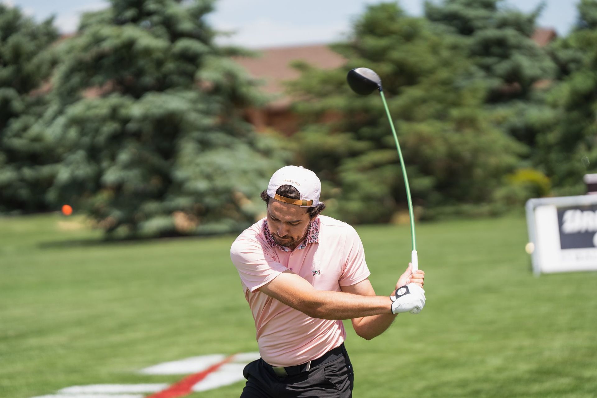 Golfer mid-swing on a green course, wearing a pink shirt and white cap, with trees in the background.