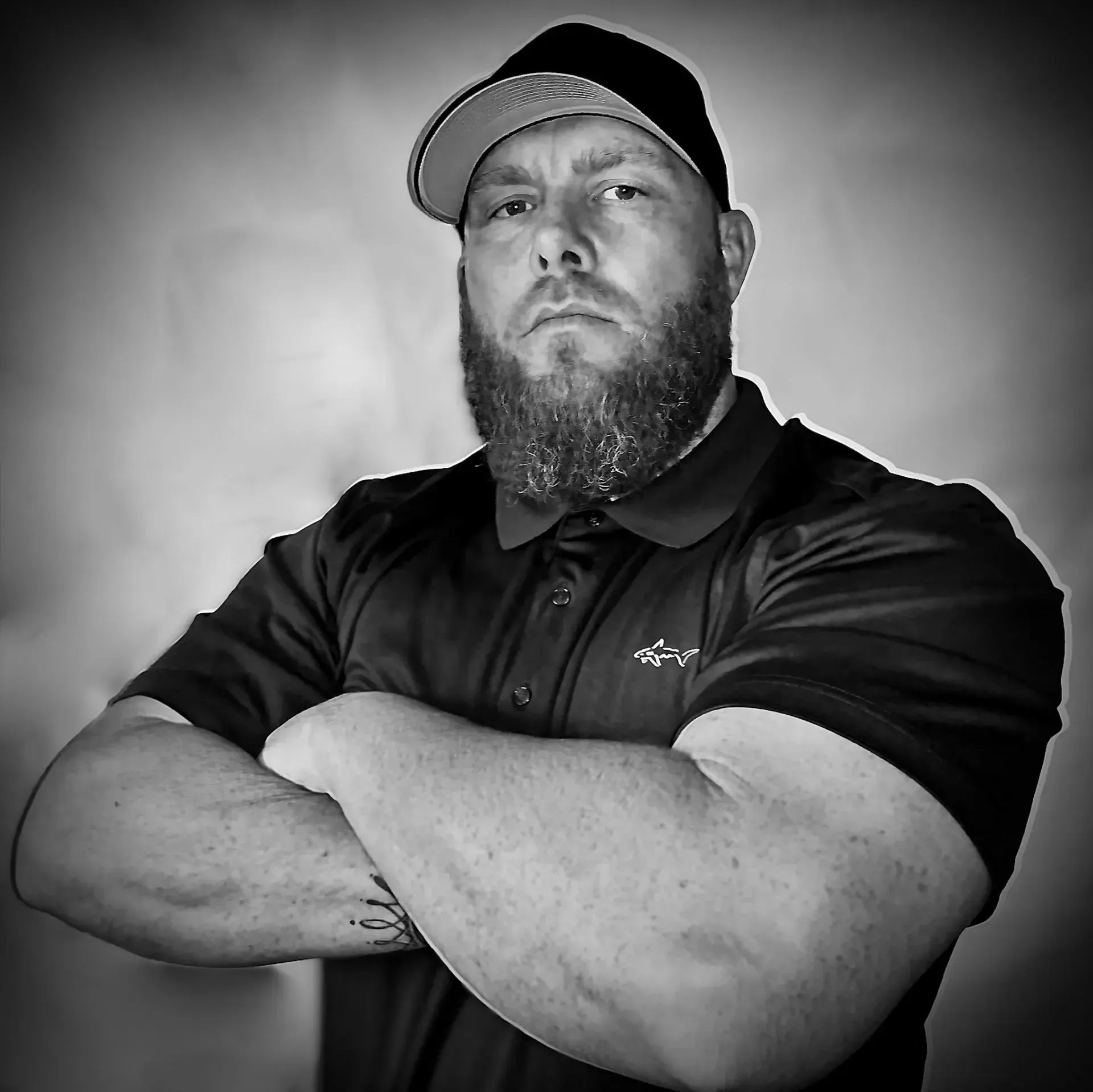 Man with a full beard, wearing a hat and polo shirt, arms crossed, looking sternly at the viewer.