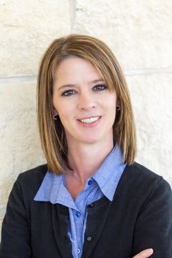 Smiling woman in a blue shirt and black cardigan against a light stone wall