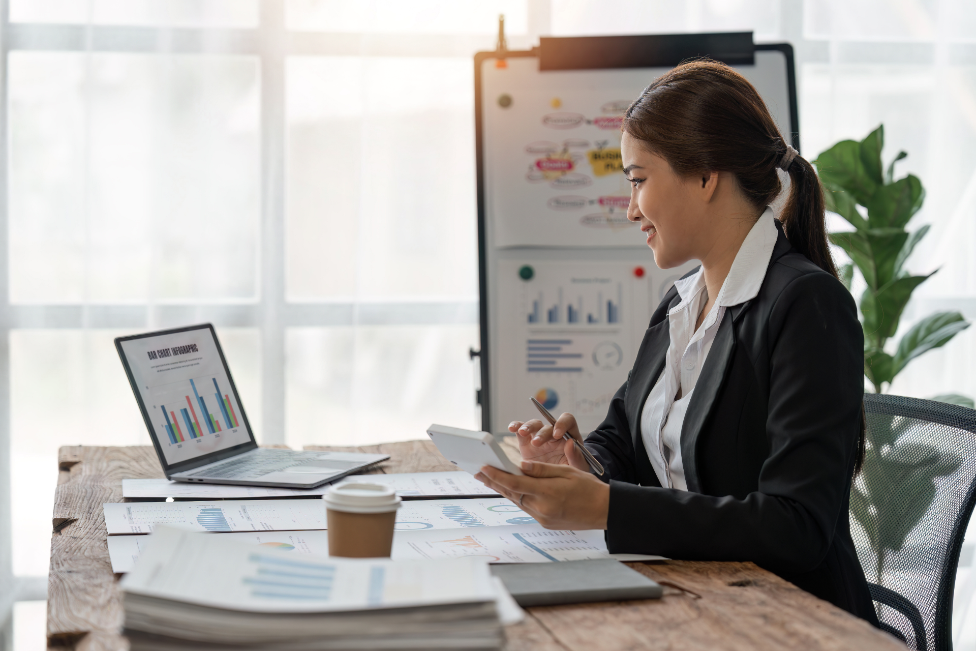 Woman in a black blazer presenting charts during a video call at a desk in a bright office