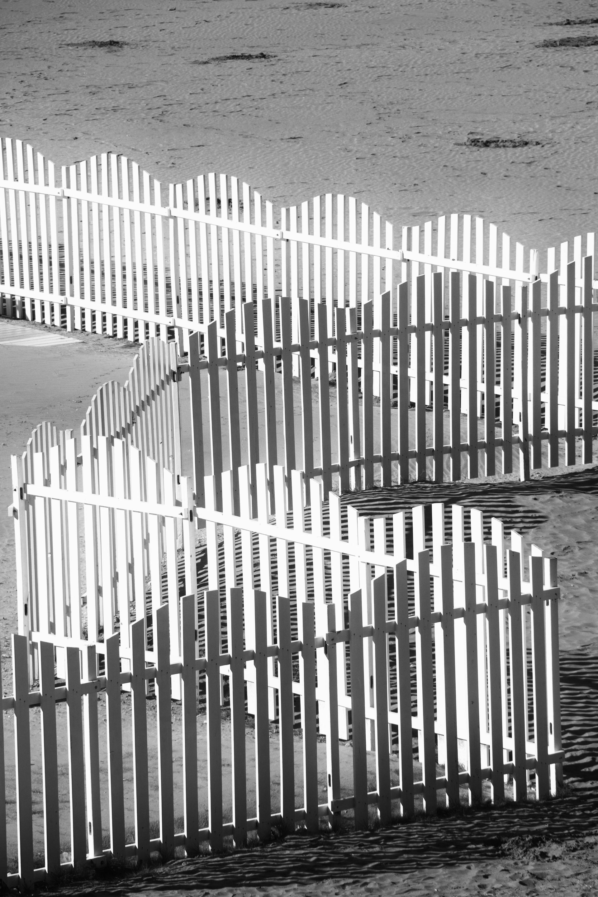 Black-and-white photo of a zigzag white picket fence on a sandy beach or shoreline.