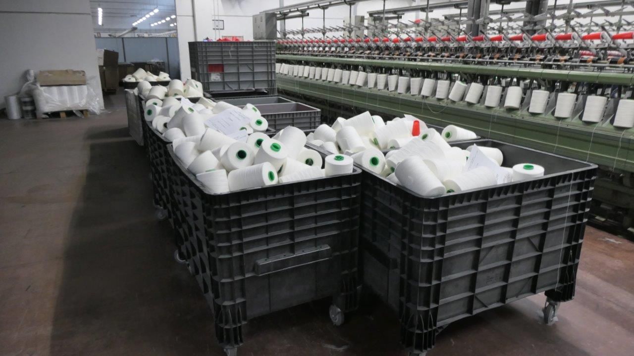 Carrelli industriali in plastica nera pieni di bobine di filato bianco all'interno di uno stabilimento di produzione tessile.