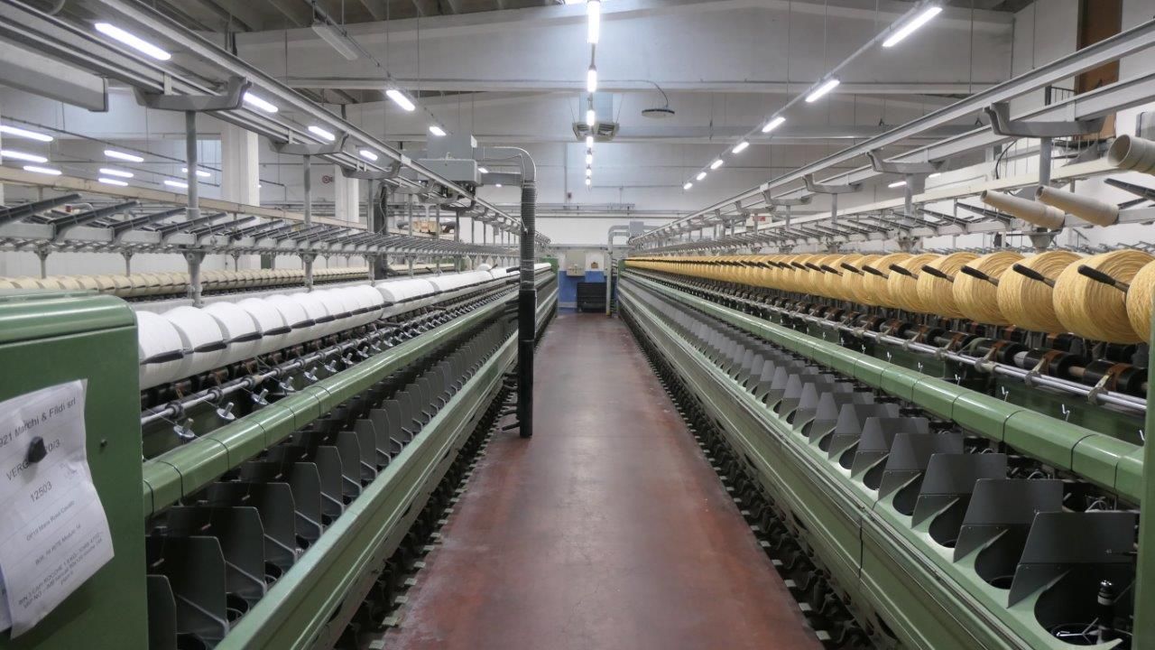 In una fabbrica tessile, filatoi industriali con file di bobine bianche e gialle, visti lungo un corridoio centrale.