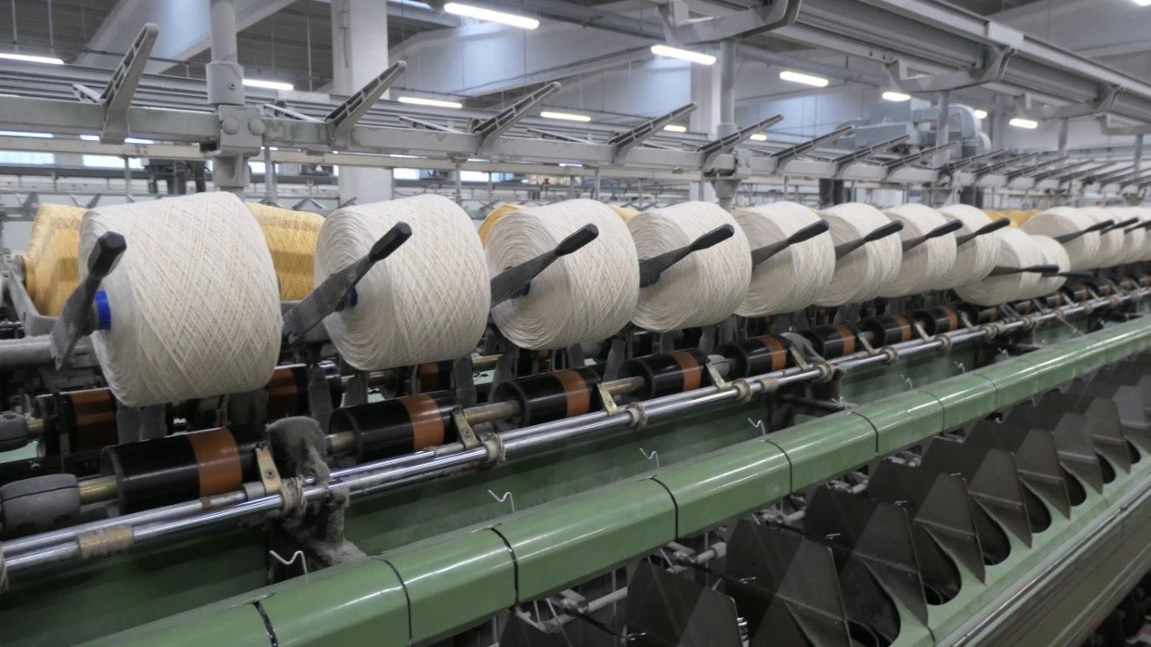 Macchinari tessili industriali con una lunga fila di rocchetti di filato bianco in un ambiente di fabbrica.