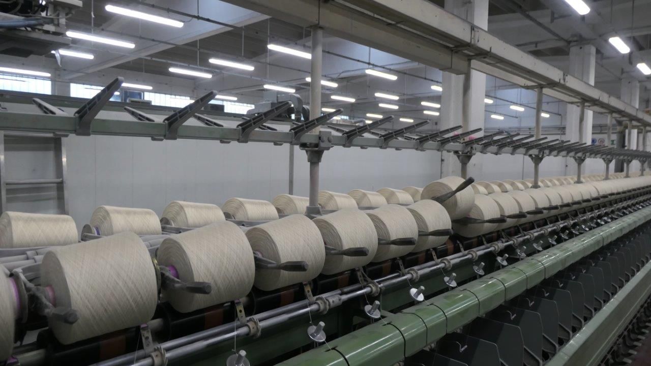File di rocchetti di filato bianco su un filatoio tessile in un ambiente industriale ben illuminato.