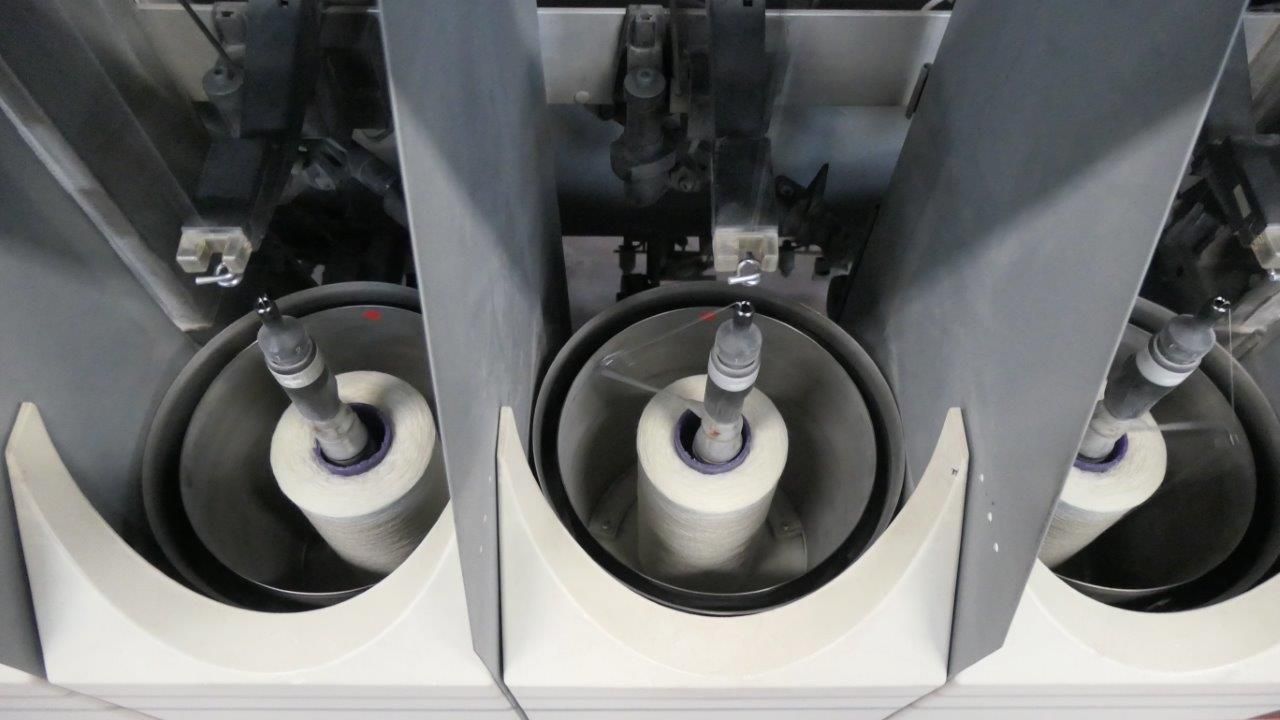 Macchina tessile industriale che avvolge filo bianco su tre bobine cilindriche alloggiate in singoli supporti metallici.