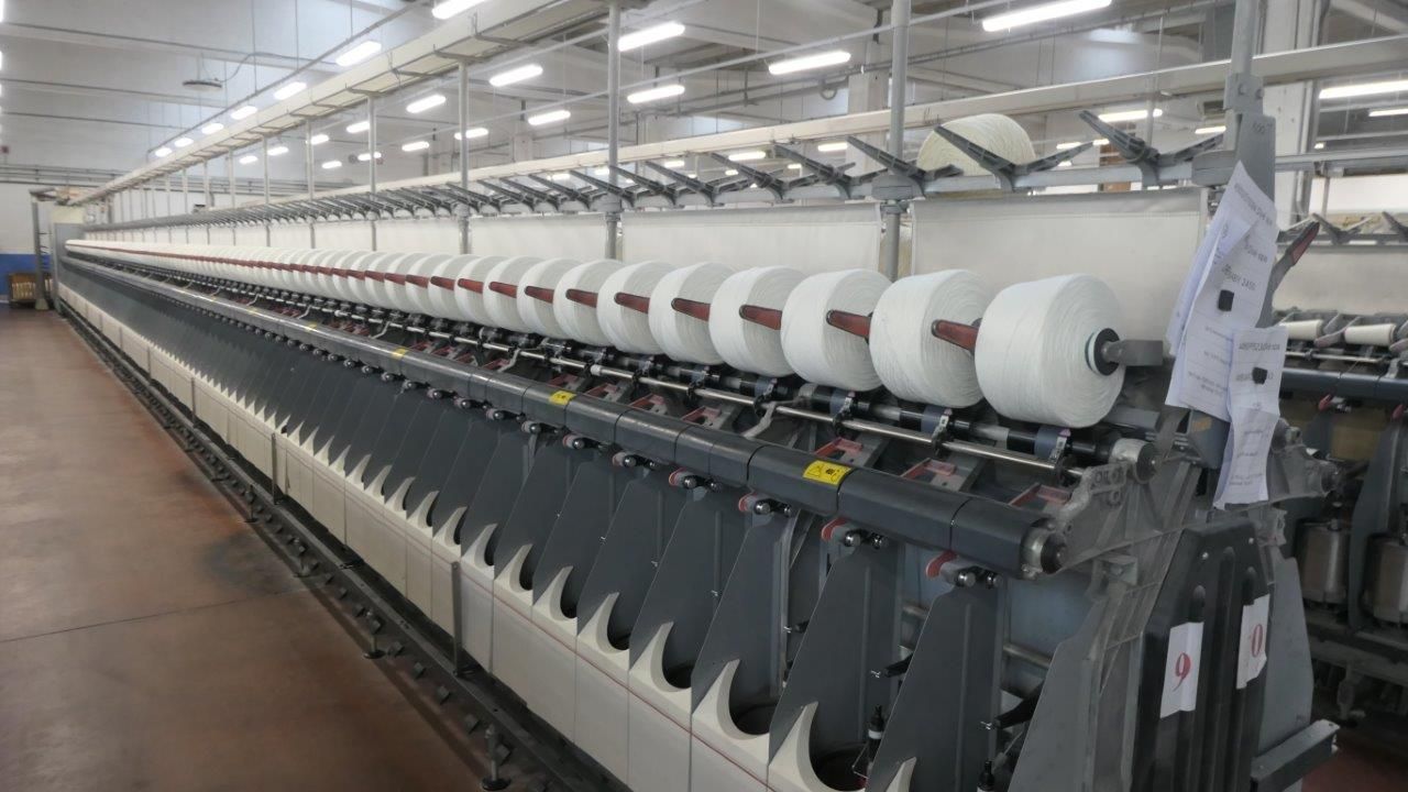 Macchina tessile industriale che avvolge filo bianco su numerose bobine in un ambiente di fabbrica.