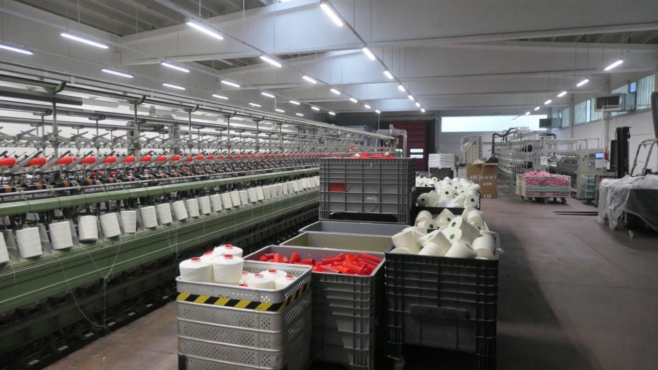 Interno di una fabbrica tessile con una lunga macchina per filare, file di bobine bianche e contenitori 