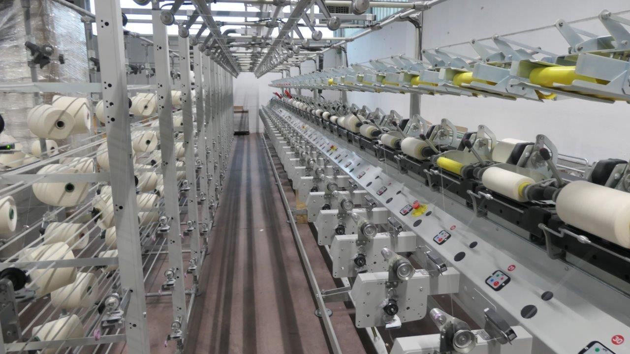 Un macchinario tessile industriale con file di bobine di filato bianco in un contesto di fabbrica.